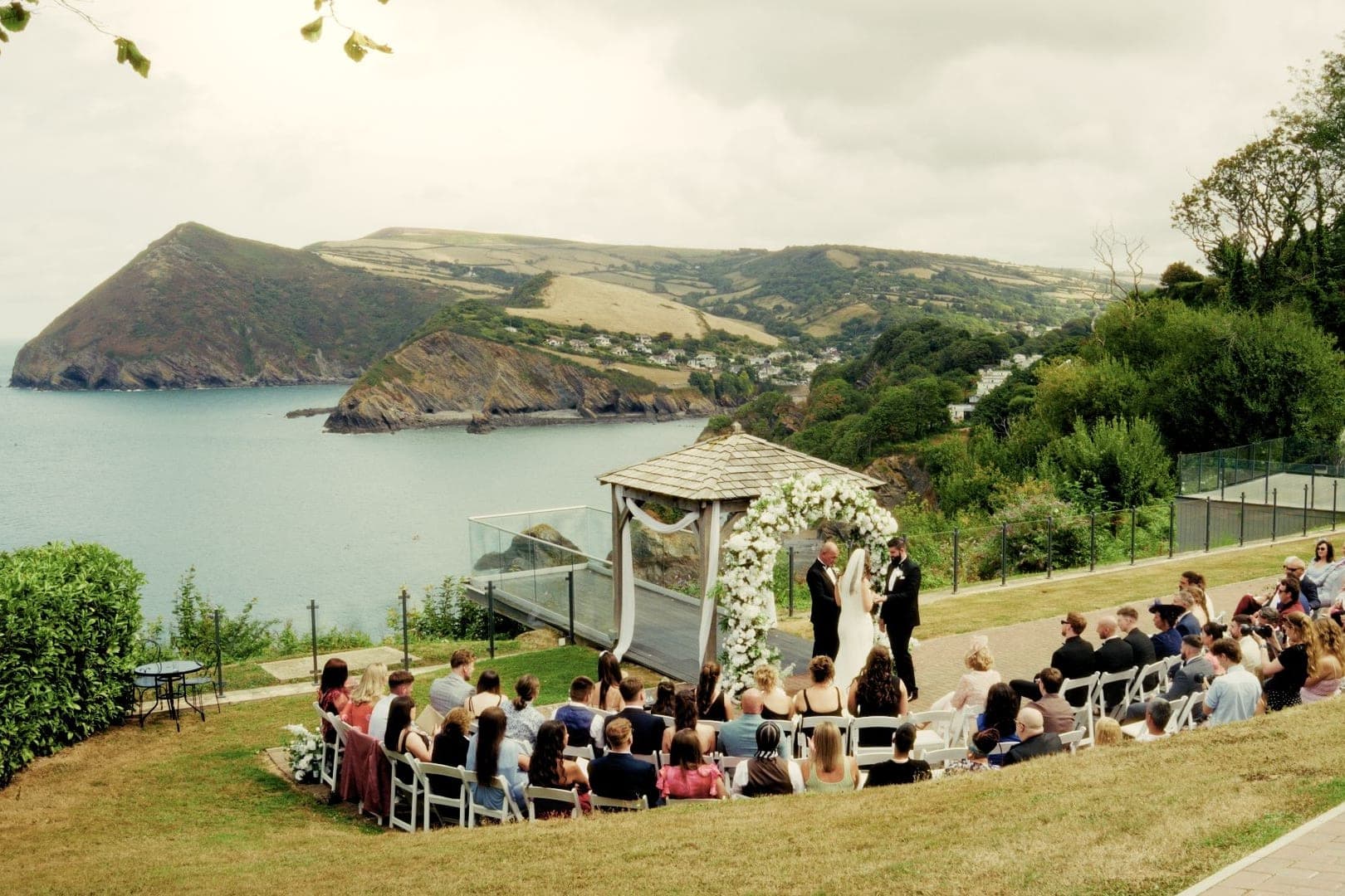 A clifftop wedding ceremony under a flower arch, guests seated on the lawn above the sea