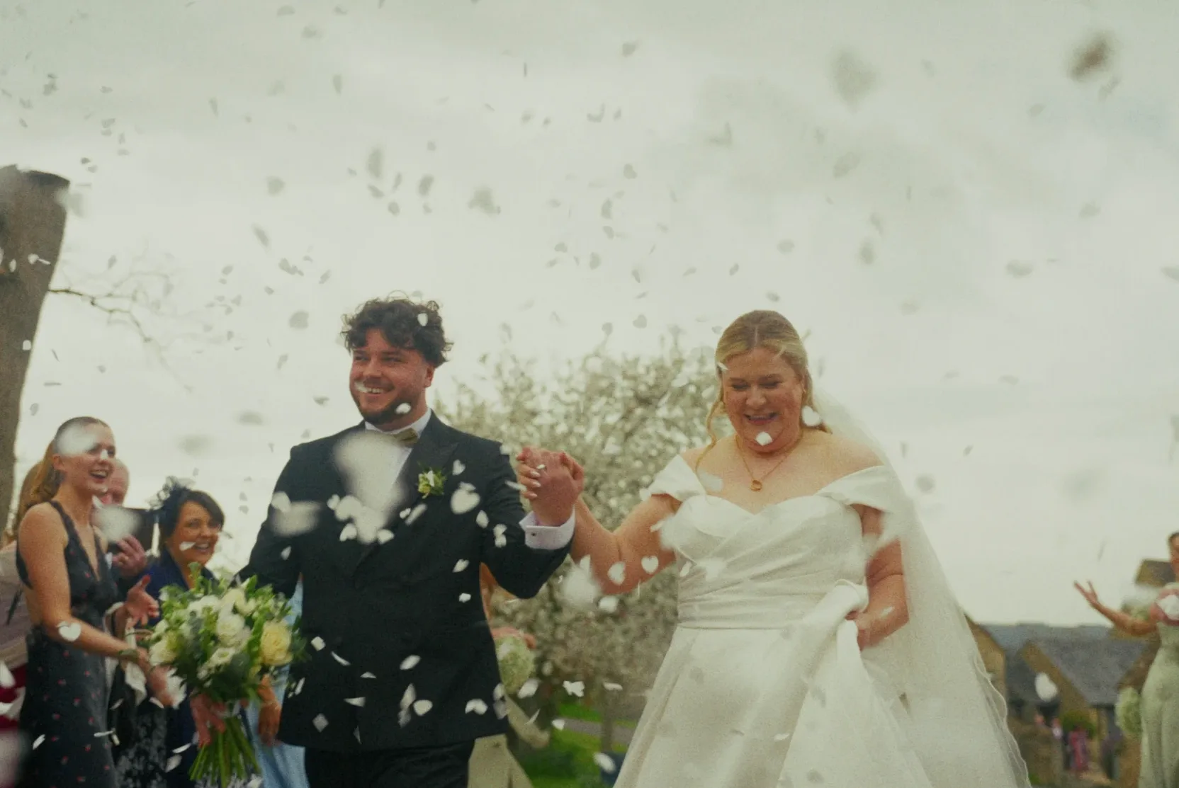 Lucy & Jack walking hand in hand through a shower of white petals, both laughing, guests cheering behind them