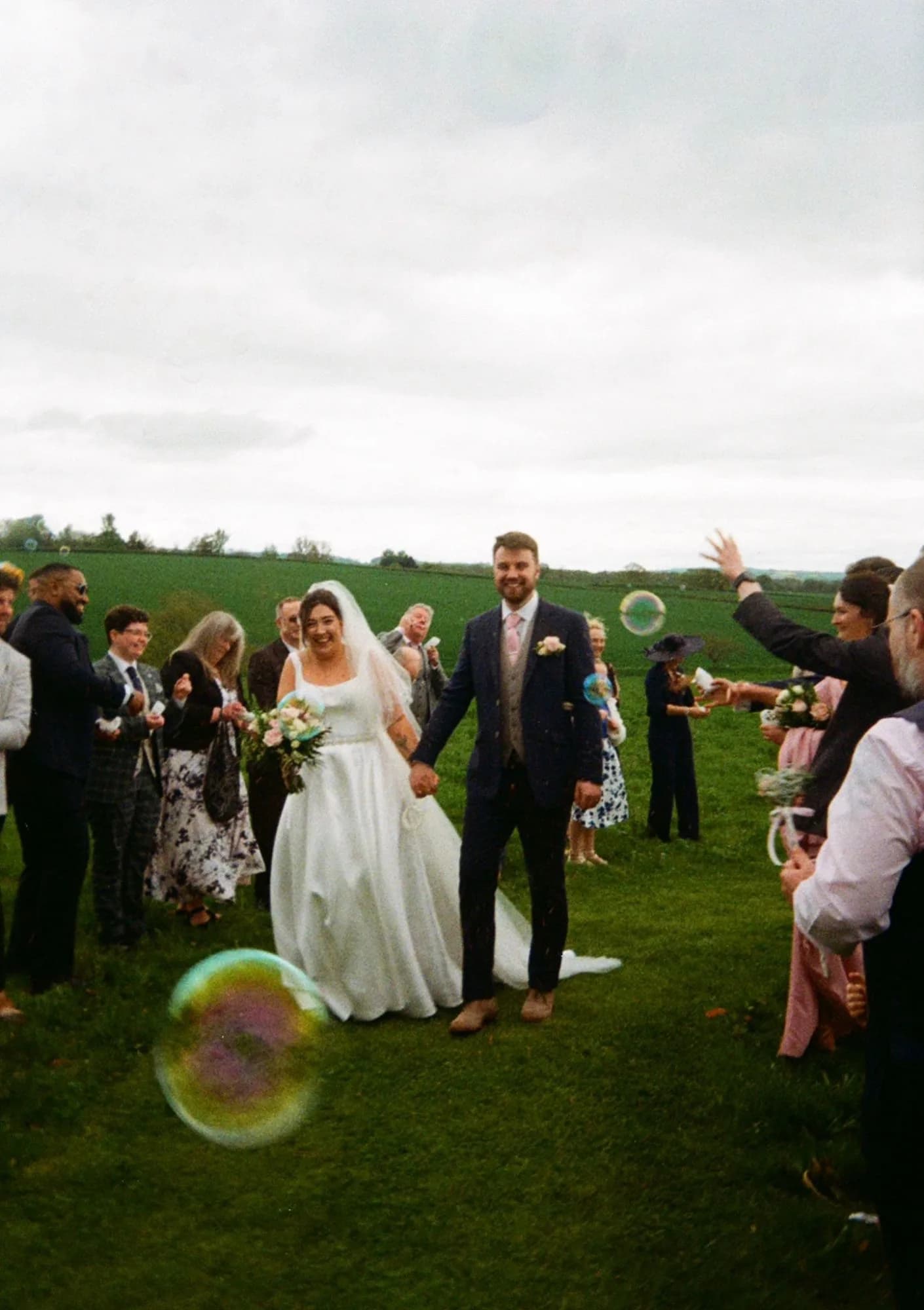 Shannon & Connor walking hand in hand across a field through bubbles, guests lining the way and cheering